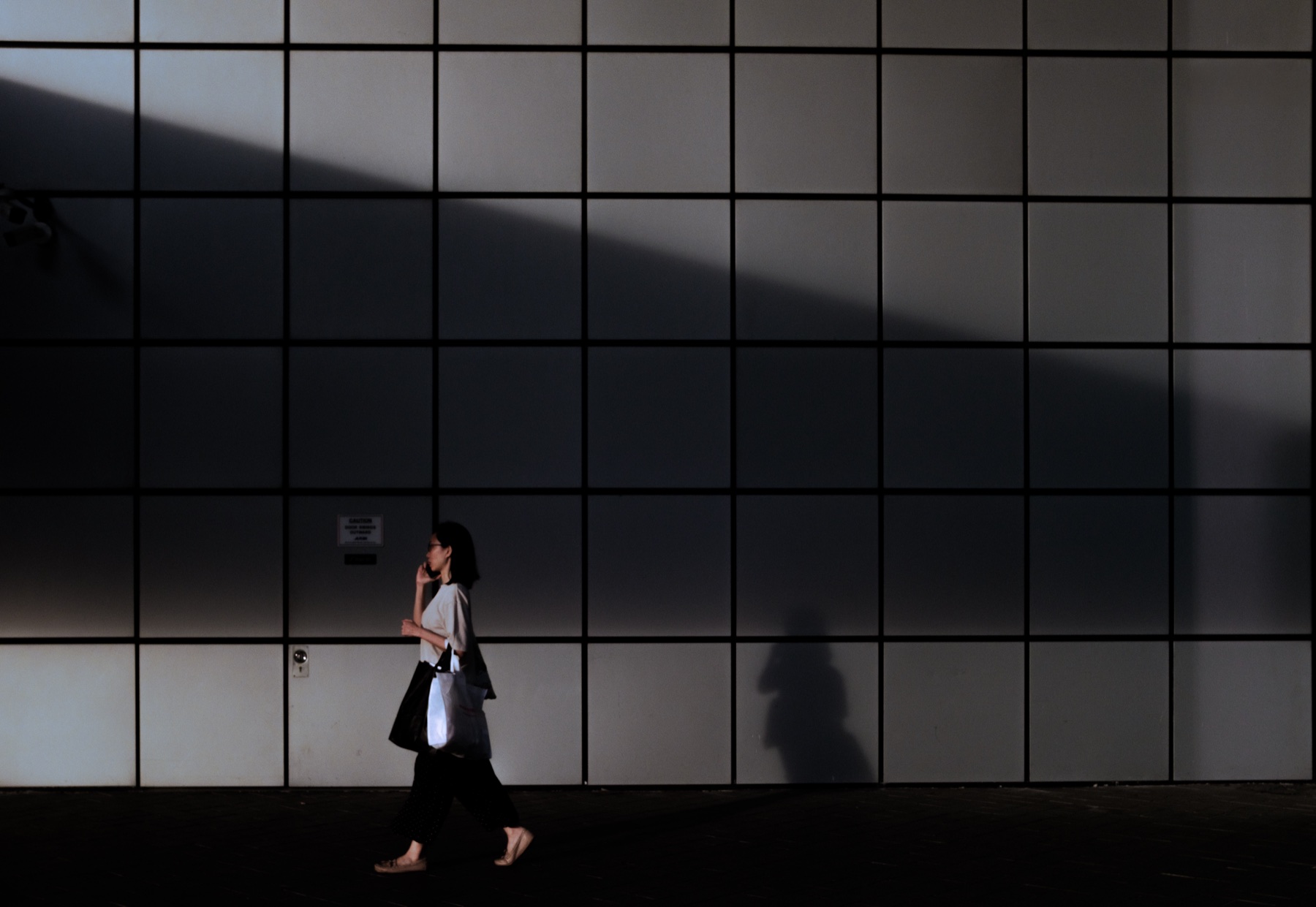 A woman walking past a grid wall, her shadow cast beside her