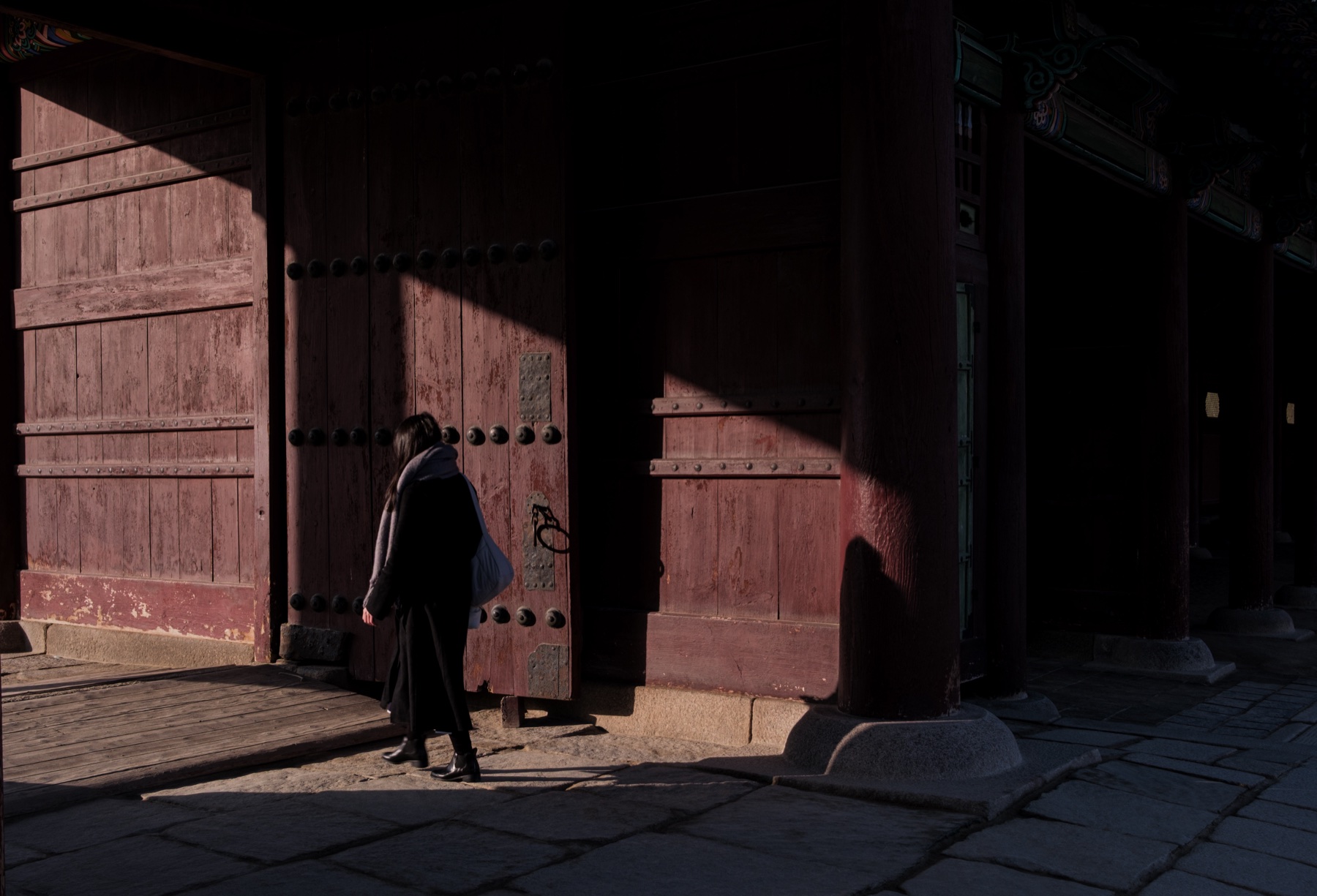 A figure in black passing tall red temple doors in raking light