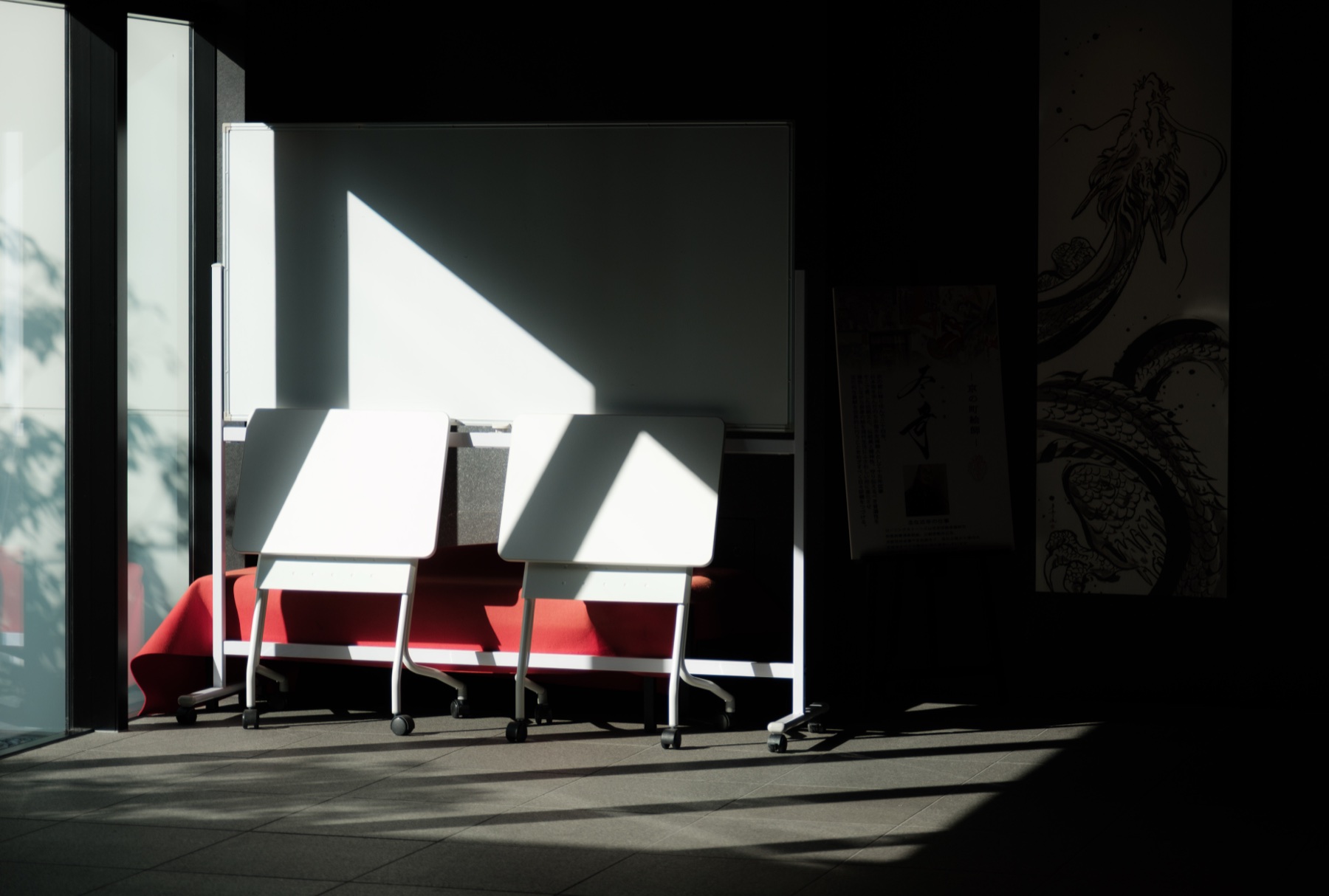 A shaft of light across white tables and a red bench beside a dragon mural