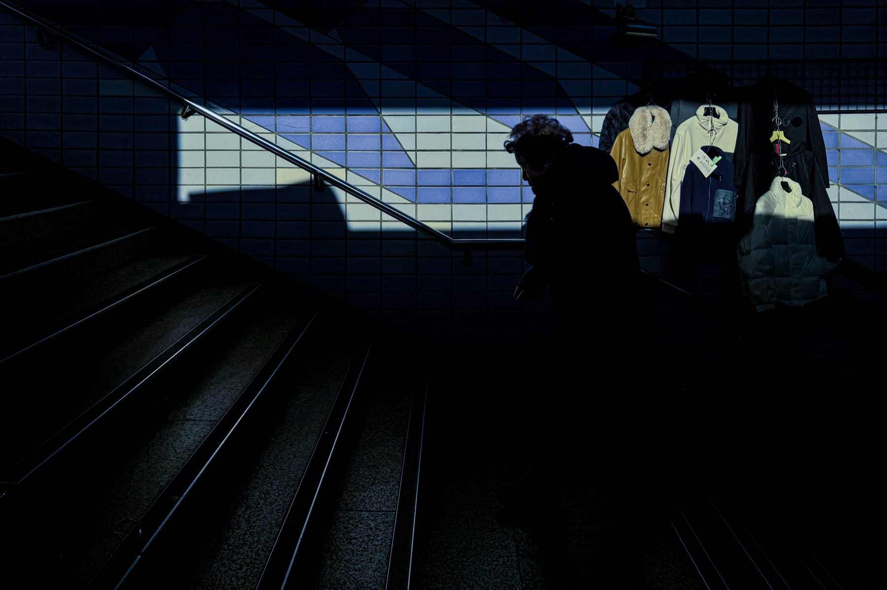 A figure descending dim subway stairs beside hanging coats